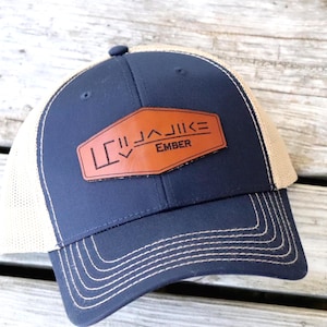 May include: A navy blue trucker hat with a tan mesh back. The front features a brown leather patch with the text "LC JAIKE EMBER" in white. The hat has a curved brim and a button on top.
