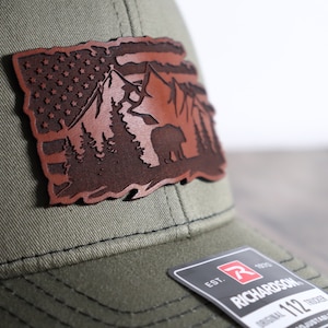 May include: A green baseball cap with a brown leather patch featuring a mountain scene with a bear, trees, and an American flag.