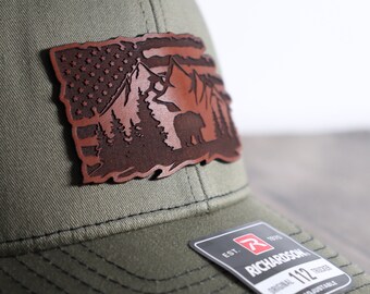 May include: A green baseball cap with a brown leather patch featuring a mountain scene with a bear, trees, and an American flag.