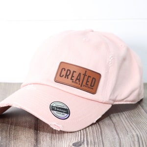 May include: Pink baseball cap with a brown leather patch that says "CREATED with a purpose" and a cross.