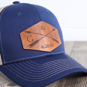 May include: A blue and beige trucker hat with a brown leather patch featuring two crossed arrows and the initials "G & L" with the date "6.20.09" underneath.