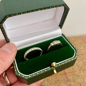 Può includere: Un portagioie verde aperto con interno bianco e chiusura dorata, contenente due anelli. Uno è in argento con piccole pietre trasparenti, e l'altro è in oro con tre pietre trasparenti. La scatola è tenuta da una persona.