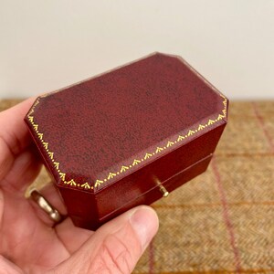 May include: A burgundy, octagonal jewelry box with gold trim. The box has a textured surface and a small gold clasp. The box is held in a hand.