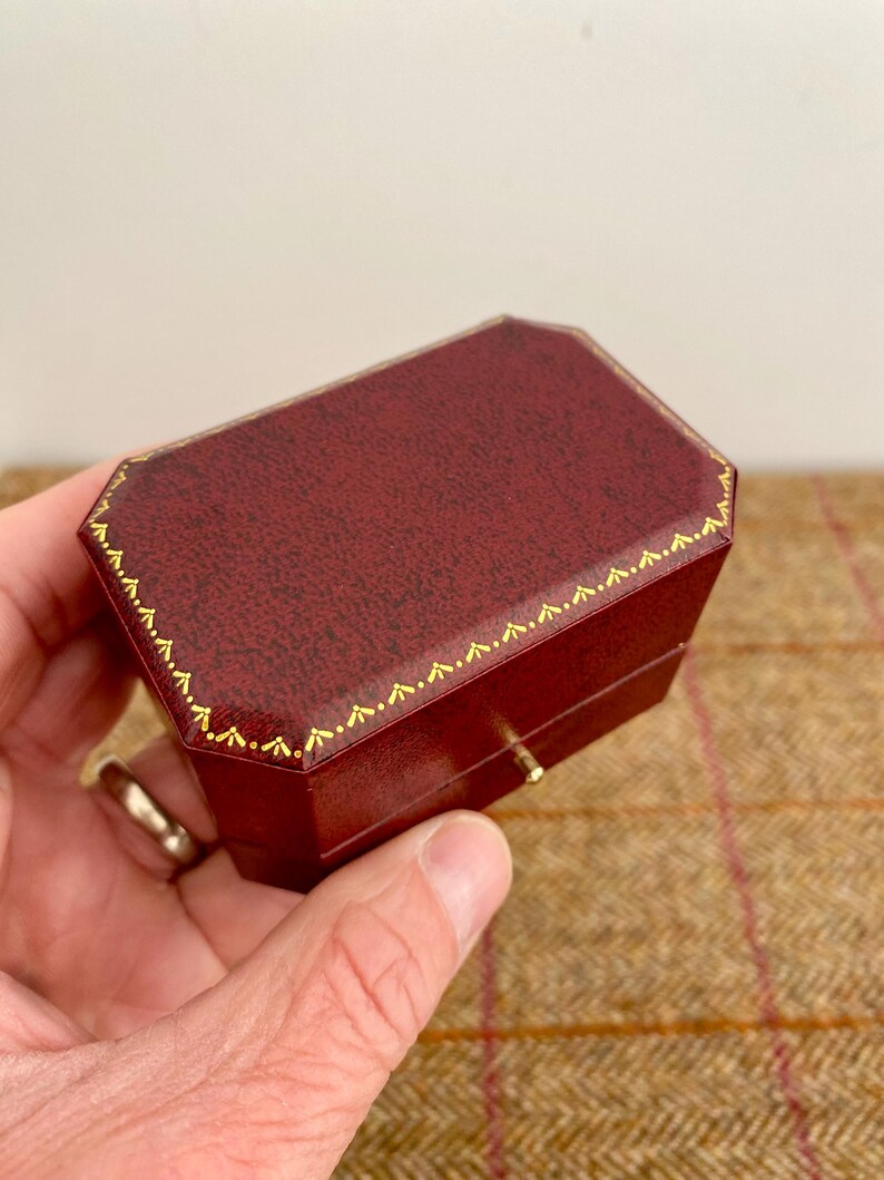 May include: A burgundy, octagonal jewelry box with gold trim. The box has a textured surface and a small gold clasp. The box is held in a hand.