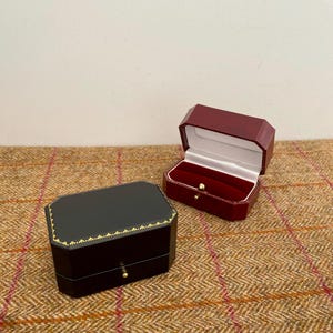 May include: Two jewelry boxes, one black and one burgundy, sit on a textured brown surface. The black box is closed with gold trim and a small gold knob. The burgundy box is open, revealing a white interior and red velvet lining.