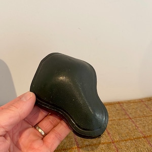 Könnte beinhalten: Ein dunkelgrünes, lederartiges Objekt mit einer gebogenen Form, möglicherweise ein Etui oder ein Behälter. Das Objekt hat eine strukturierte Oberfläche und scheint geschlossen zu sein. Der Hintergrund ist neutral.