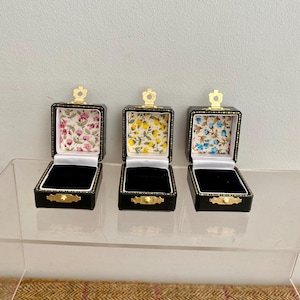 Peut inclure: Trois boîtes à bagues carrées noires avec des fermoirs et des charnières en métal doré. Chaque boîte présente une base intérieure en velours noir et un couvercle doublé de tissu avec des motifs floraux : roses roses, fleurs jaunes et bleues.