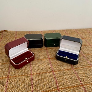 Pode incluir: Quatro pequenas caixas de joias abertas em bordô, preto e verde, com interiores de veludo branco ou azul. Cada caixa tem um fecho dourado. As caixas estão sobre uma superfície estampada marrom e vermelha.