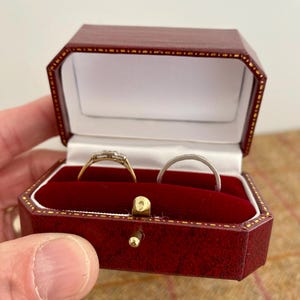 May include: A red leatherette jewelry box with gold trim, open to reveal two rings on a red velvet interior. One ring is gold with a square design, the other is a silver band.