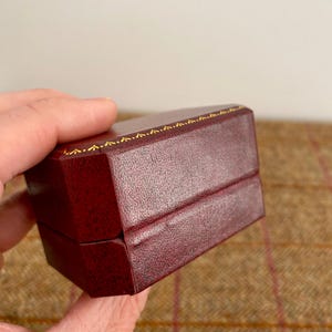 May include: A burgundy jewelry box with a textured surface. The box has a rectangular shape with a hinged lid. The edges are trimmed with a gold-colored decorative pattern. The box is held in a person's hand.