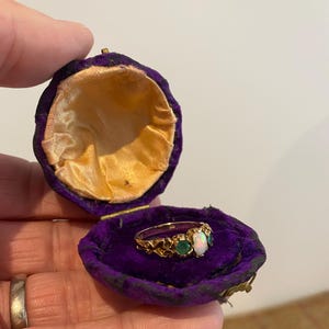 May include: A gold ring featuring an opal and two emeralds, set in a decorative band, is displayed in an open purple velvet ring box with a gold satin lining. The ring box is open.