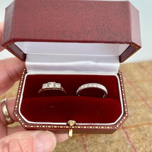 May include: A red jewelry box with a gold trim, open to reveal two rings. One ring is gold with three square diamonds, and the other is a silver band with small diamonds. The box is lined with red velvet.