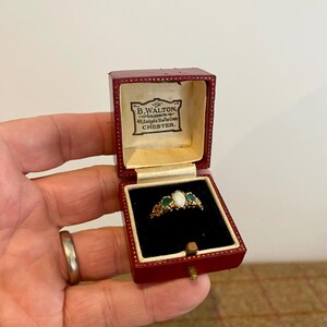 May include: A gold ring featuring a central oval opal flanked by two green stones, presented in a deep red hinged box. The interior lid displays the text B.WALTON, GOLDSMITH, 45, Eastgate St, The Cross, CHESTER. The ring rests on a black velvet base.