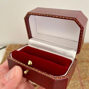 May include: A burgundy, octagonal ring box with a red velvet interior. The box is open, revealing two slots for rings. Gold-colored accents trim the edges and the clasp. The interior is lined with white satin.