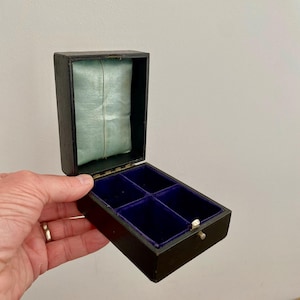 May include: Open black jewellery box with a blue velvet interior and a light blue satin pillow. The box is divided into four compartments. The box is held open by a hand.