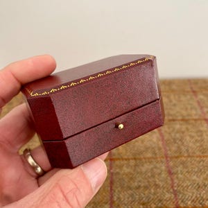 May include: A burgundy-colored, octagonal jewelry box with a gold-colored trim. The box has a small, gold-colored knob on the front. The box is held in a person's hand.