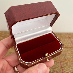 May include: Open burgundy ring box with a white satin interior and a red velvet insert. The box has a gold-colored clasp and a textured exterior. The box is octagonal in shape.