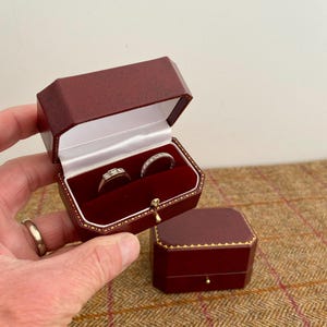 May include: Two rings displayed in a burgundy leather ring box with a gold clasp. The box is open, revealing a white interior and a red velvet lining. A second, closed box is also visible.