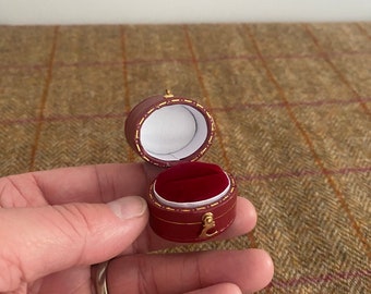 May include: A small, red ring box with a gold trim and a velvet interior. The box is open and the velvet lining is visible.