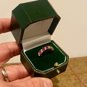 Pode incluir: Um anel de ouro com pedras preciosas vermelhas e transparentes, apresentado numa caixa de joias verde forrada a veludo. A caixa é verde escuro com acabamentos dourados e um fecho dourado.