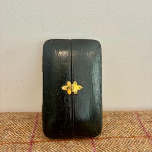 May include: A rectangular black wooden box with a glossy finish, featuring a central gold-coloured metal latch. The box is hinged and has rounded corners, presenting a smooth, dark surface with visible wood grain texture under the polished coating.