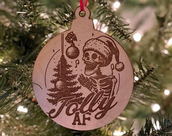 Pode incluir: Um enfeite de Natal de madeira em forma de círculo com um esqueleto vestindo um chapéu de Papai Noel segurando uma árvore de Natal. O enfeite é gravado com o texto "Jolly AF".