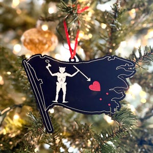 May include: Black and red Christmas bauble shaped like a pirate flag with a skull and crossbones design. The flag has a red heart and three red dots on it.