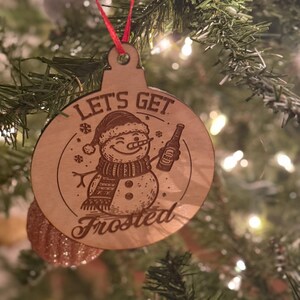 May include: Wooden Christmas ornament with a snowman design. The ornament features the text "Let's Get Frosted" and a snowman holding a bottle. The ornament is hanging on a Christmas tree with a red ribbon.