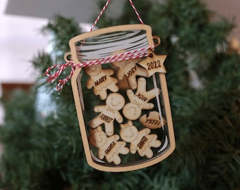Puede incluir: Un adorno navideño de madera con forma de tarro de cristal con un lazo de cuerda rojo y blanco. Dentro del tarro hay ocho figuras de pan de jengibre con nombres escritos en ellas. El adorno también tiene una estrella con el año 2022 en ella.