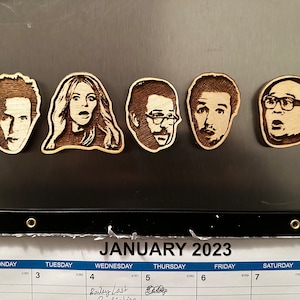 May include: Five wooden magnets with engraved portraits of people with surprised expressions. The magnets are attached to a dark surface. The calendar below shows the month of January 2023.
