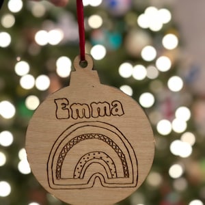 Puede incluir: Un adorno navideño de madera con un diseño de arcoíris y el nombre "Emma" grabado en él.