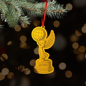 May include: A gold-colored flat ornament shaped like a winged figure holding a globe above their head, standing on a cylindrical base. The base features the engraved text NICK RAMSEY DIRECTOR OF PHOTOGRAPHY. A red ribbon is attached for hanging.