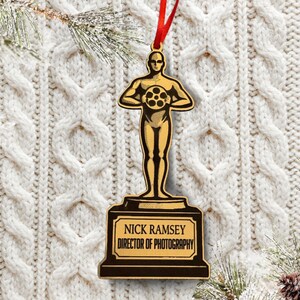 May include: Wooden Christmas ornament shaped like an Oscar statuette, with a red ribbon for hanging. The statuette holds a film reel and is mounted on a base with the text "NICK RAMSEY DIRECTOR OF PHOTOGRAPHY".