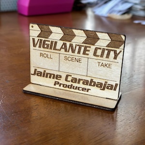 May include: Wooden film clapperboard with the text "VIGILANTE CITY" and the name "Jaime Carabajal, Producer." The clapperboard has a dark brown top and a light brown base, standing on a wooden surface.