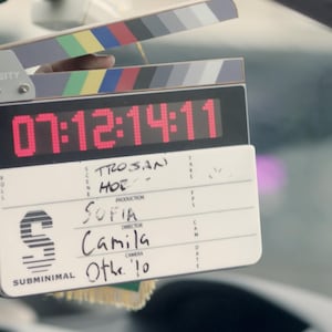 Könnte beinhalten: Eine weiße Filmklappe mit einem schwarzen Griff und einer digitalen Uhr, die 07:12:14:11 anzeigt. Die Klappe trägt die Aufschrift "TROSAN HOE" sowie den Produktionsnamen "SOFIA", den Namen des Regisseurs "Camila" und den Namen des Kameramannes "Othe' 10".