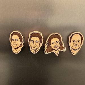 Könnte beinhalten: Vier Holzmagnete mit den Gesichtern der Hauptfiguren aus der Fernsehserie Seinfeld. Die Magnete befinden sich in einer Reihe auf einem silbernen Kühlschrank.