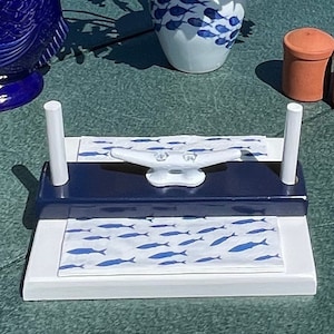 May include: A white and blue nautical themed napkin holder with a white ceramic cleat on top. The holder has two white posts and a dark blue base. The napkin holder is sitting on a green surface.