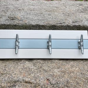 May include: A rectangular wooden wall-mounted coat rack with four silver-colored boat cleat hooks. The rack is painted white with a light blue stripe in the center. The hooks are evenly spaced across the board.