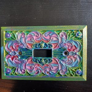 Puede incluir: Cubierta de interruptor de luz decorativa con un diseño multicolor. De forma rectangular, presenta una abertura central y orificios para tornillos. El diseño floral es rosa, azul y verde, enmarcado por un borde verde.