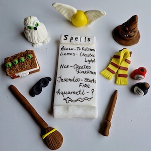 Può includere: Un set di topper per torte a tema Harry Potter, tra cui un boccino d'oro, un gufo bianco, un cappello marrone, una sciarpa rosso e gialla, una maschera nera, una bacchetta marrone, una scopa marrone, un libro con occhi verdi e un rotolo con il testo "Spells Accio-To Retrieve Lumos-Creates Light Nox-Creates Darkness Insendio-Starts Fire Aquameti-?".
