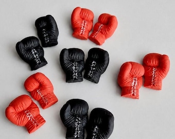 May include: A collection of miniature boxing glove charms in solid red and black colors. Each glove features detailed molded textures, including a laced front closure and a padded wrist section, arranged in a scattered pattern.