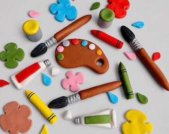 May include: A collection of clay art supplies including two paintbrushes with brown handles and black bristles, a brown paint palette with colourful dots, two paint tubes, two crayons, and various scattered clay shapes in red, blue, yellow, green, and pink.