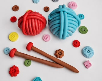 Könnte beinhalten: Ein roter und ein blauer Wollknäuel werden mit zwei braunen Stricknadeln und verschiedenen bunten Knöpfen gezeigt. Die Knöpfe sind in den Farbtönen Blau, Rosa, Gelb, Braun und Rot gehalten. Das Bild ist aus der Vogelperspektive aufgenommen.