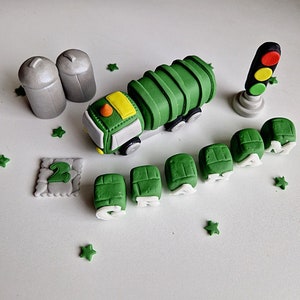 May include: A collection of green and white fondant cake decorations, including a garbage truck, trash cans, a traffic light, and the number 2. Green star-shaped sprinkles are scattered around the objects. The garbage truck has yellow and white accents.