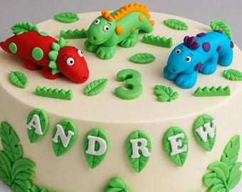 May include: A round cake with smooth off-white frosting, decorated with three colorful dinosaur figures in red, green, and blue. The top features a green number 3 and small green leaves. The side displays the name ANDREW in white letters.