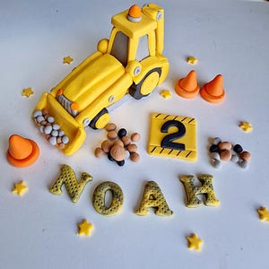 Może przedstawiać: Żółty plastikowy pojazd budowlany z łyżką, numerem 2 i imieniem Noah napisanym żółtymi literami na białym torcie.