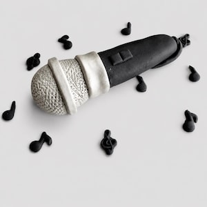 Pode incluir: Um microfone preto e prateado com uma cabeça texturizada e uma faixa branca. Notas musicais pretas circundam o microfone em um fundo branco. O microfone é um item decorativo, possivelmente um topo de bolo ou um artesanato similar.