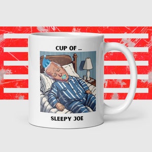 Puede incluir: Taza de café de cerámica blanca con una ilustración de dibujos animados de una persona durmiendo en la cama. El texto "CUP OF... SLEEPY JOE" está impreso en la taza.