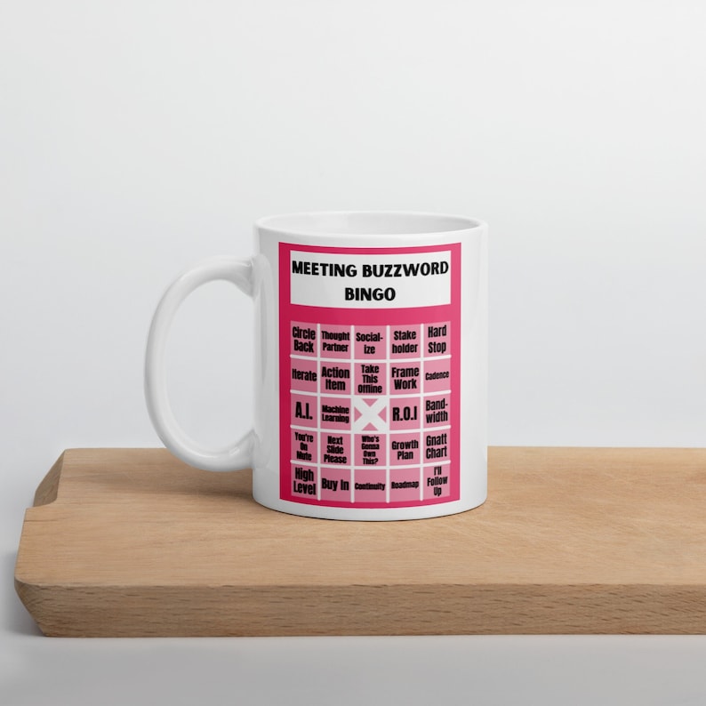 Puede incluir: Taza de cer&aacute;mica blanca con un dise&ntilde;o rosa. El dise&ntilde;o presenta una tarjeta de bingo con palabras clave de reuni&oacute;n como "Circle Back", "Thought Partner", "Socialize", "Stake Holder", "Hard Stop", "Iterate", "Action Item", "Take This Offline", "Frame Work", "Cadence", "A.I.", "Machine Learning", "R.O.J", "Bandwidth", "You're on Mute", "Next Slide Please", "Who's Gonna Take This?", "Growth Plan", "Gnatt Chart", "High Level", "Buy In", "Continually", "Roadmap", "I'll Follow Up"