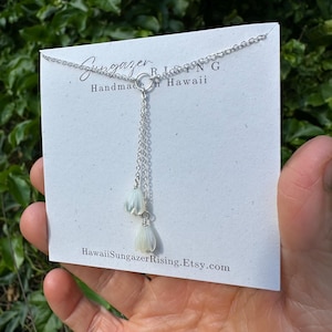 May include: A delicate silver necklace with a double-strand pendant featuring two white, flower-shaped charms. The necklace is displayed on a white card with the text "Sungazer Rising Handmade in Hawaii" and a website address.